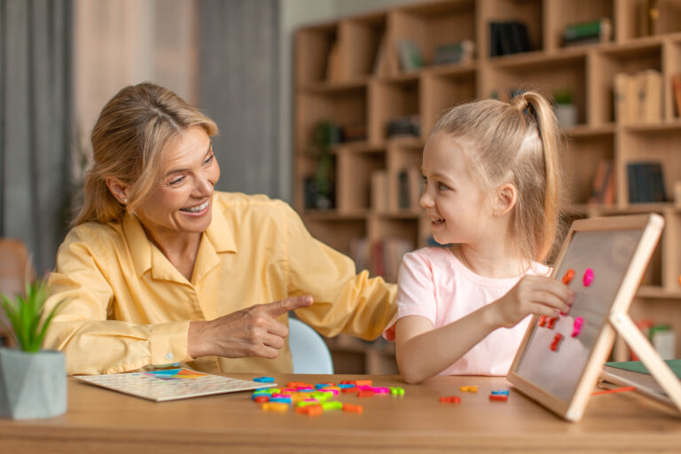 Excited woman speech therapist teaching adorable little girl alphabet, making words from plastic letters on whiteboard