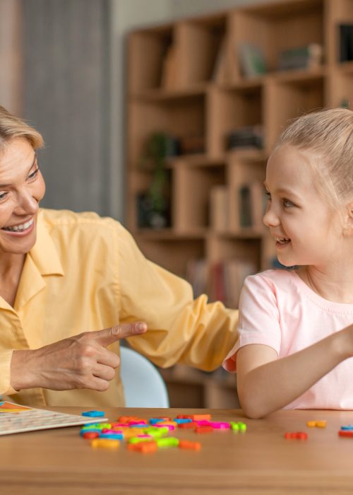 Excited woman speech therapist teaching adorable little girl alphabet, making words from plastic letters on whiteboard. Speech-language therapy for children concept