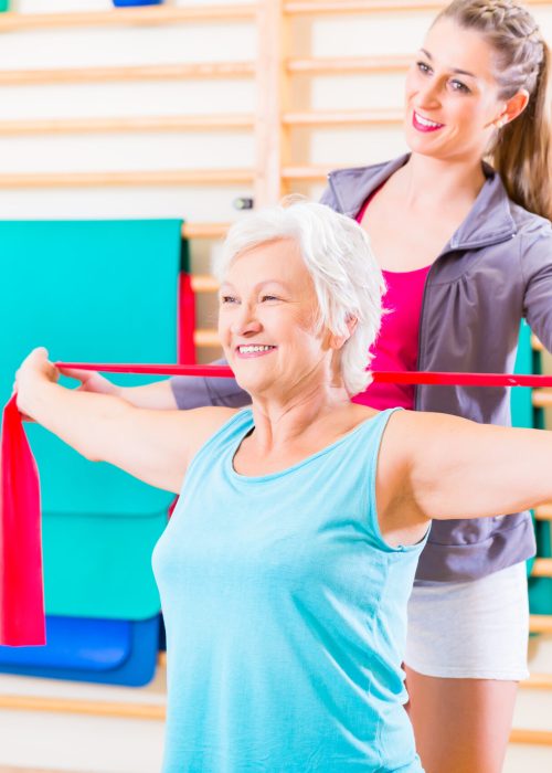 Senior woman with stretch band in fitness gym being coached by personal trainer